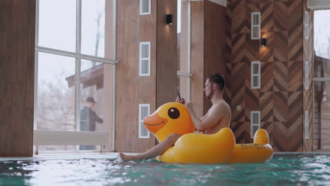 fun-and-rest-in-water-park-adult-man-is-sitting-on-inflatable-duck-in-swimming-pool-and-using-smartphone